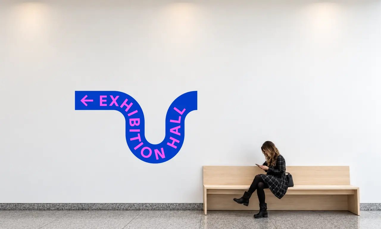 Blue curved wayfinding sign reading "Exhibition Hall" with an arrow, mounted on a white wall above a person sitting on a wooden bench