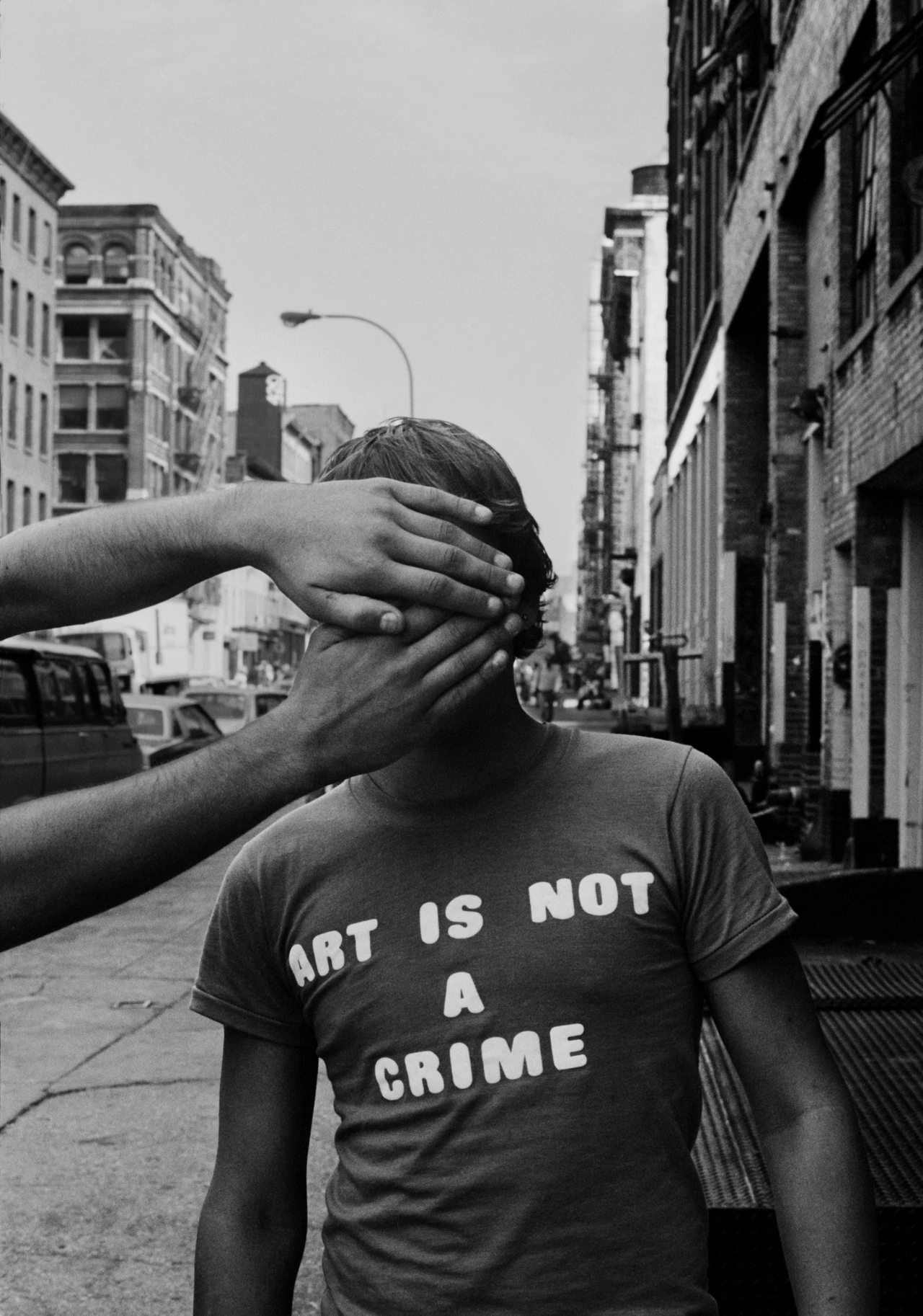 Person wearing "Art is Not a Crime" t-shirt has their face covered by hands on a city street lined with brick buildings