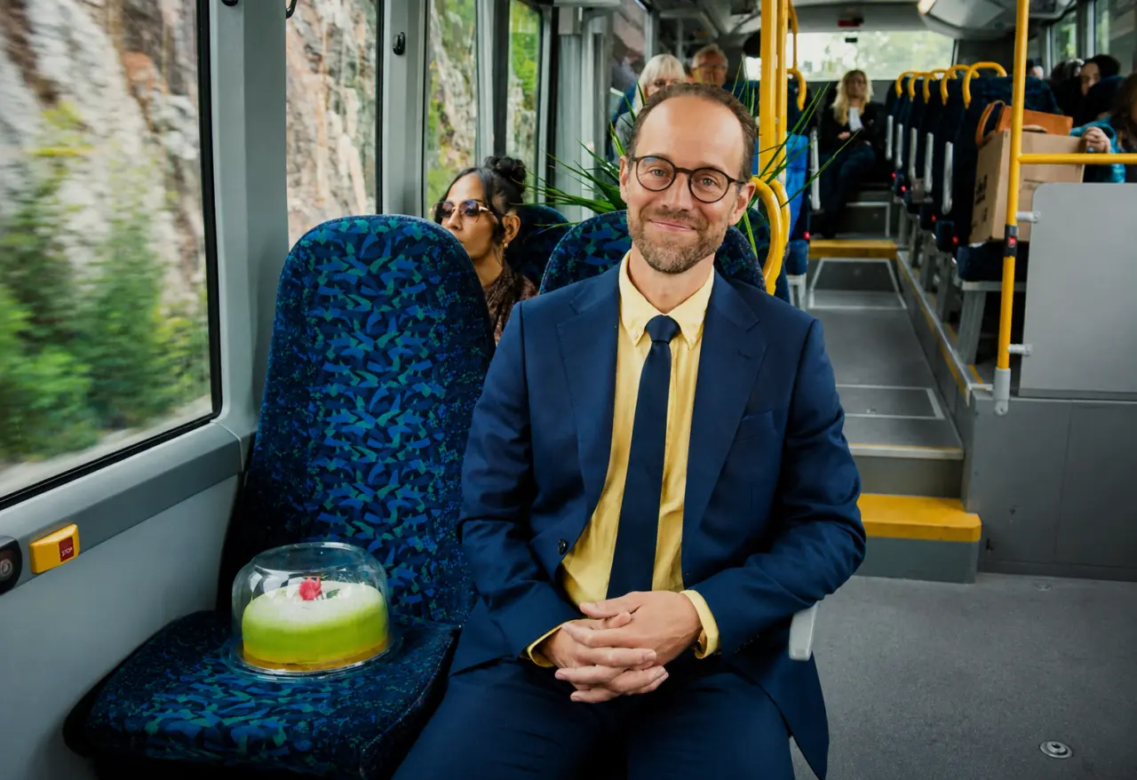 Man in navy suit and yellow tie sitting on a bus seat holding a cup of green juice, with other passengers visible behind him
