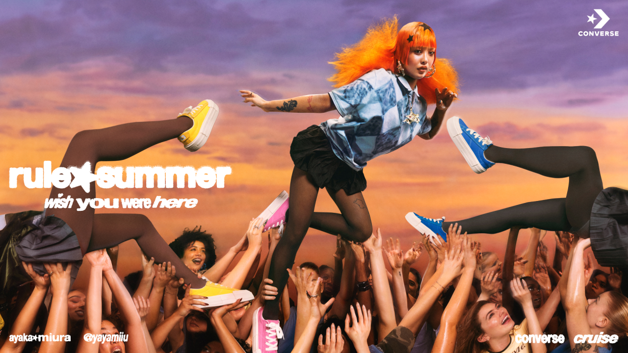 Person with bright orange hair crowd-surfing above an audience against a sunset sky, wearing blue Converse trainers, for Converse's Wish You Were Here campaign