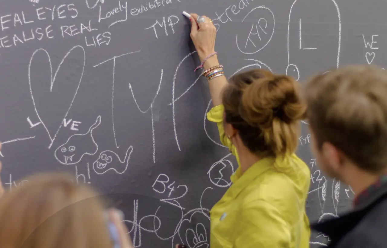 Child in yellow shirt writing on a chalkboard covered with drawings and handwritten text during a collaborative creative activity