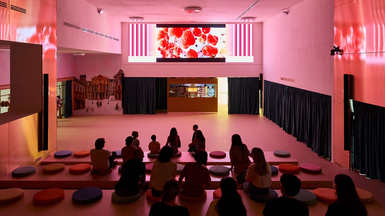 Visitors seated on cushions in a pink-lit room with a digital screen displaying red flowers above them at Leone's Happiness Factory installation
