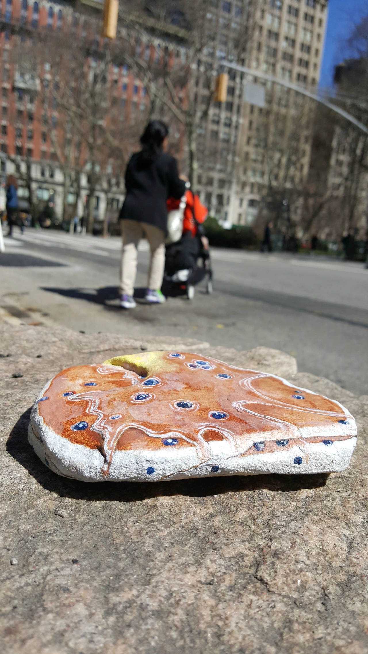 A painted rock depicting a slice of berry-studded cake sits on a city pavement with blurred pedestrians and tall buildings behind it