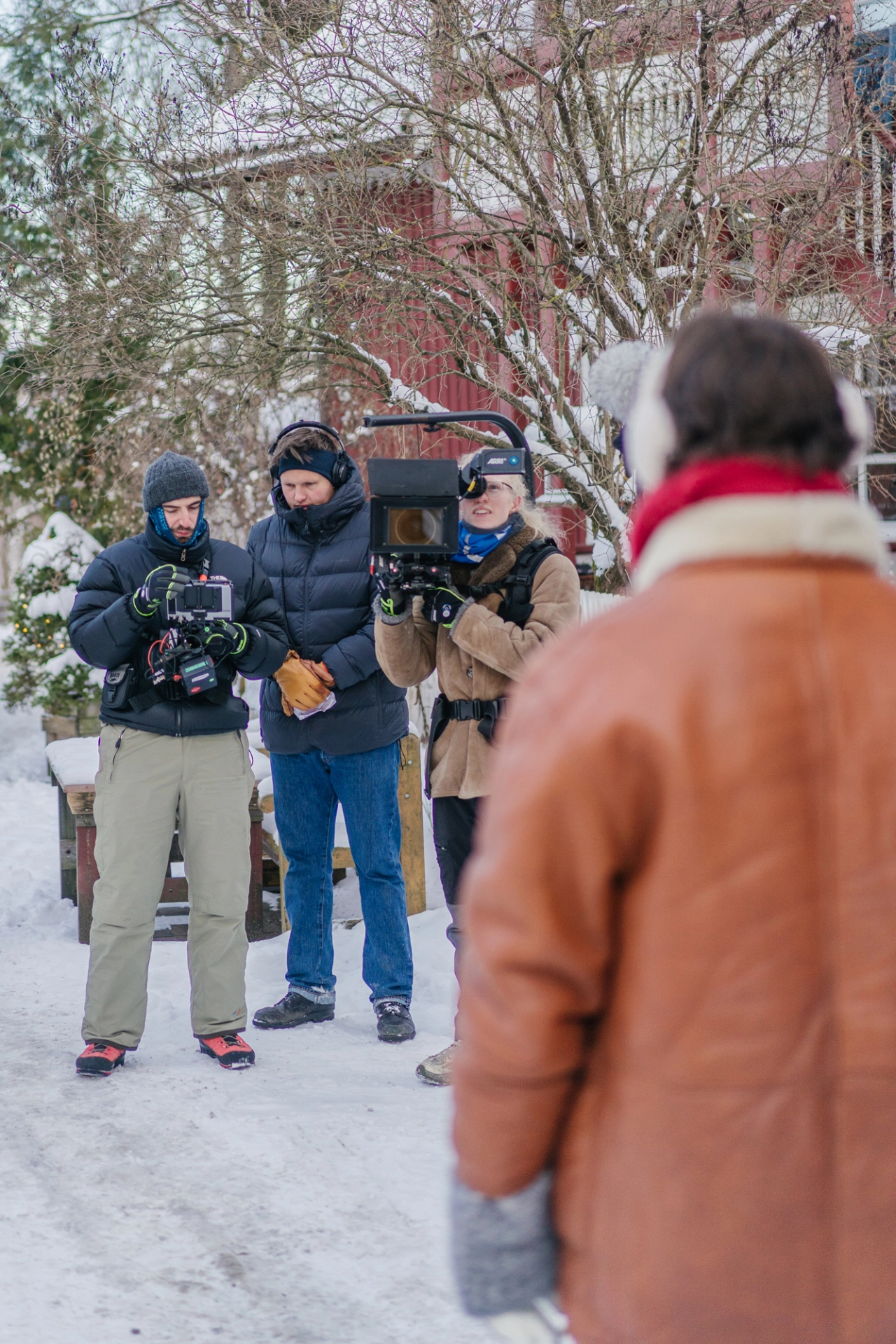 Film crew with camera and audio equipment filming a scene with a person in an orange jacket on a snowy street lined with bare trees and brick buildings