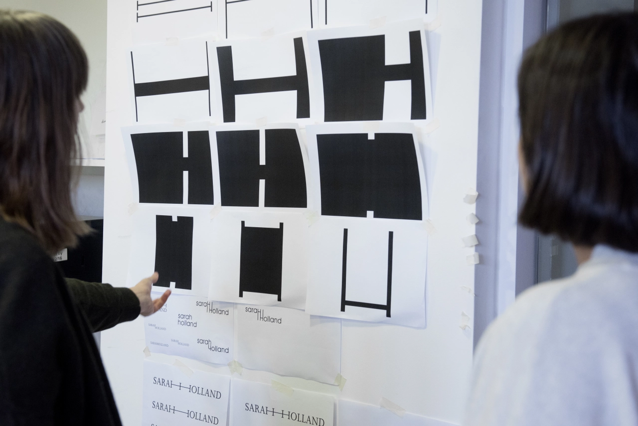 Two people examine a wall display of black and white graphic design layouts and typography samples arranged in a grid format