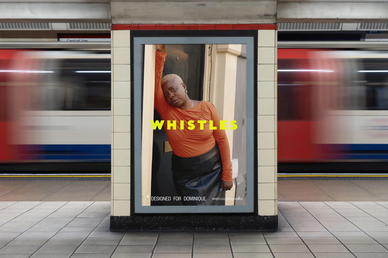 Man in orange shirt wearing headphones leans against doorframe in Whistles campaign poster displayed in London Underground station with passing train