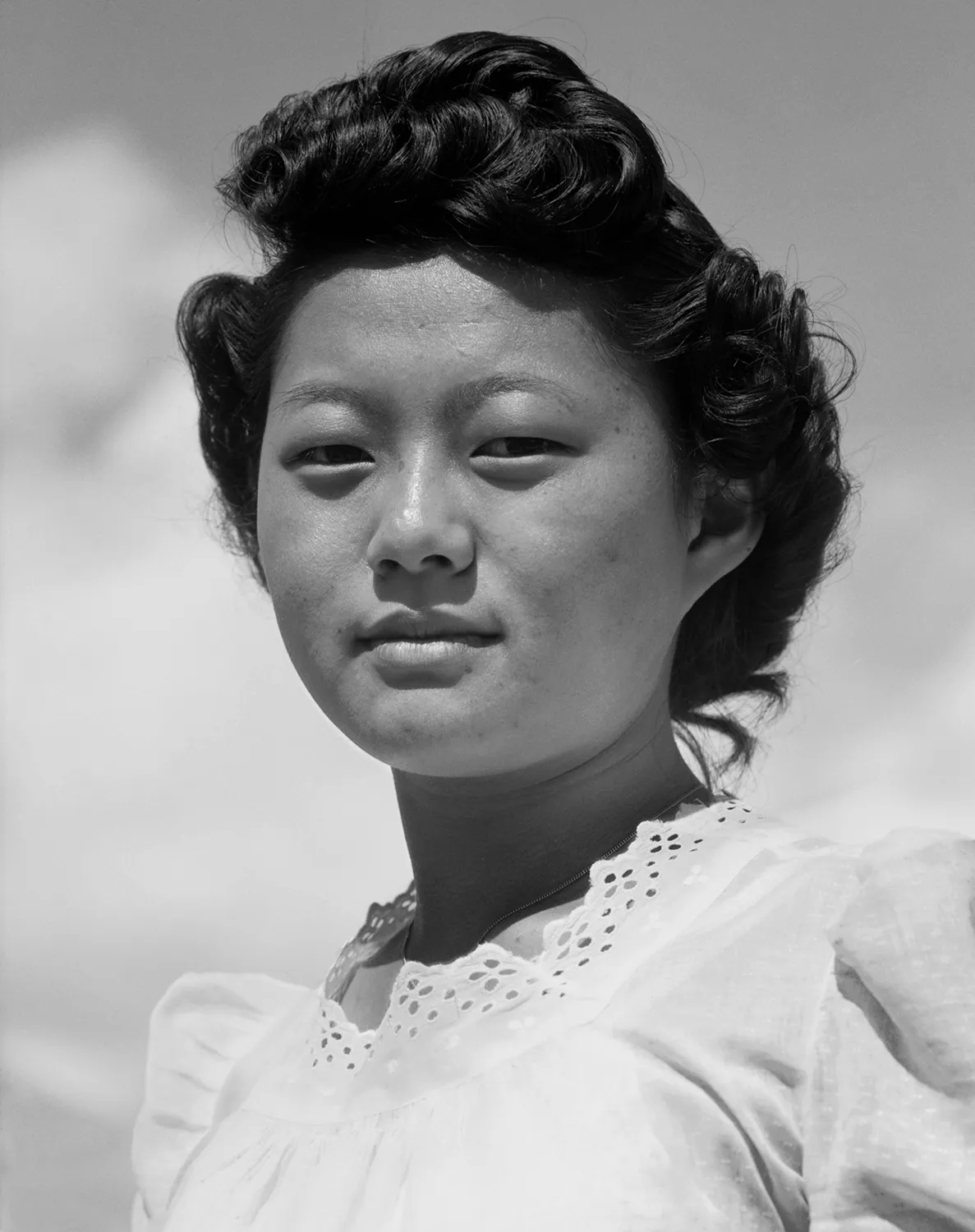 Black and white portrait photograph of a Japanese American woman with dark wavy hair and a white blouse with eyelet detailing, looking directly at the camera