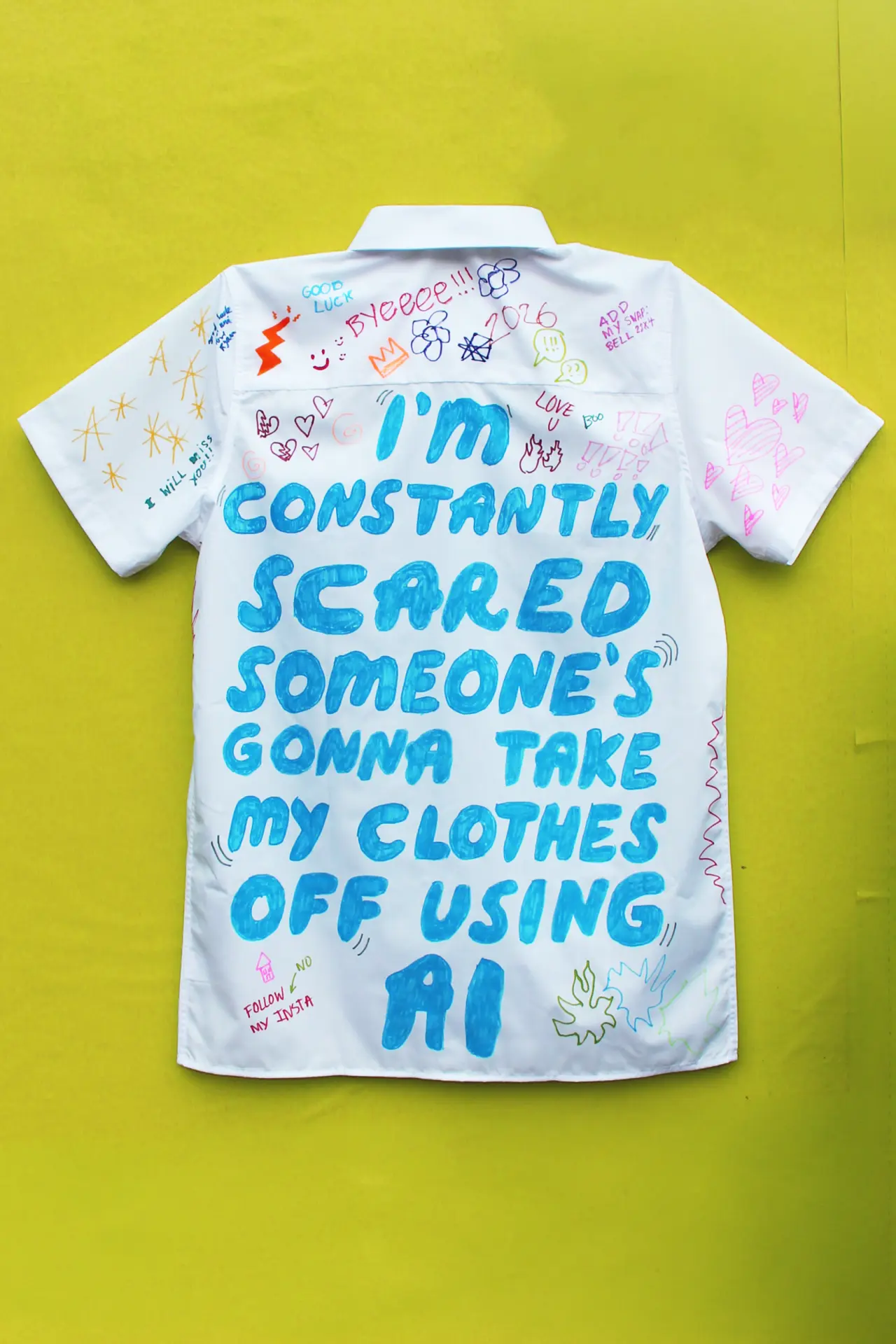 White shirt with blue handwritten text reading "I'm constantly scared someone's gonna take my clothes off using AI" surrounded by colourful doodles on yellow background