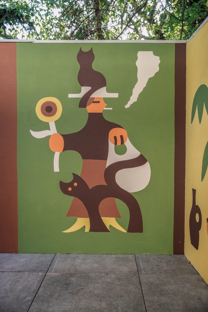 Abstract mural featuring stylised figures in brown, orange and cream on a green background, with a seated cat, a standing figure holding a circular object, and a black cat at the base