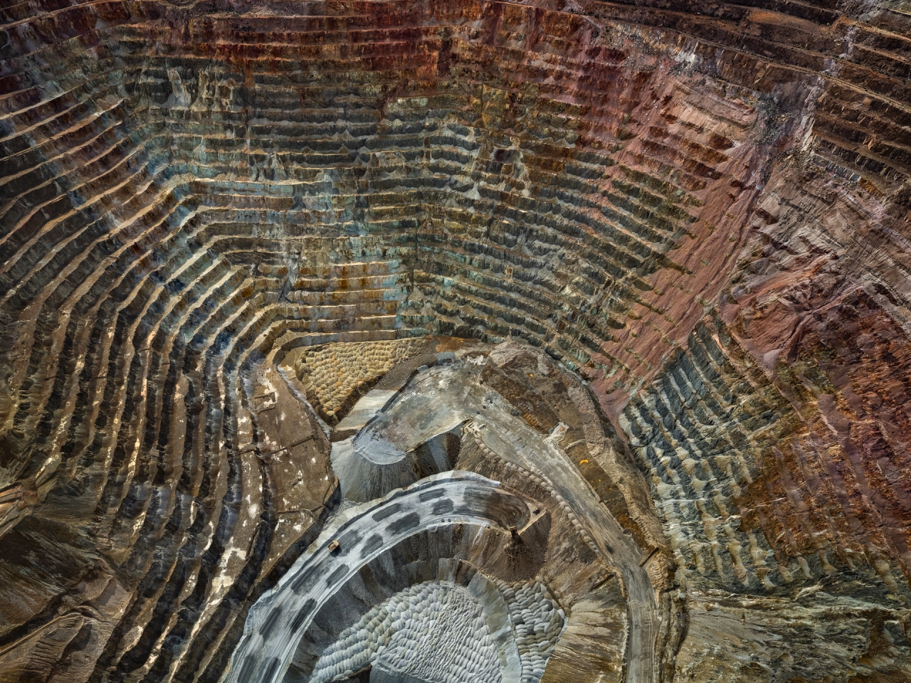 Aerial view of a vast open-pit mine with concentric terraced layers in shades of green, brown and rust, revealing the scale of mineral extraction
