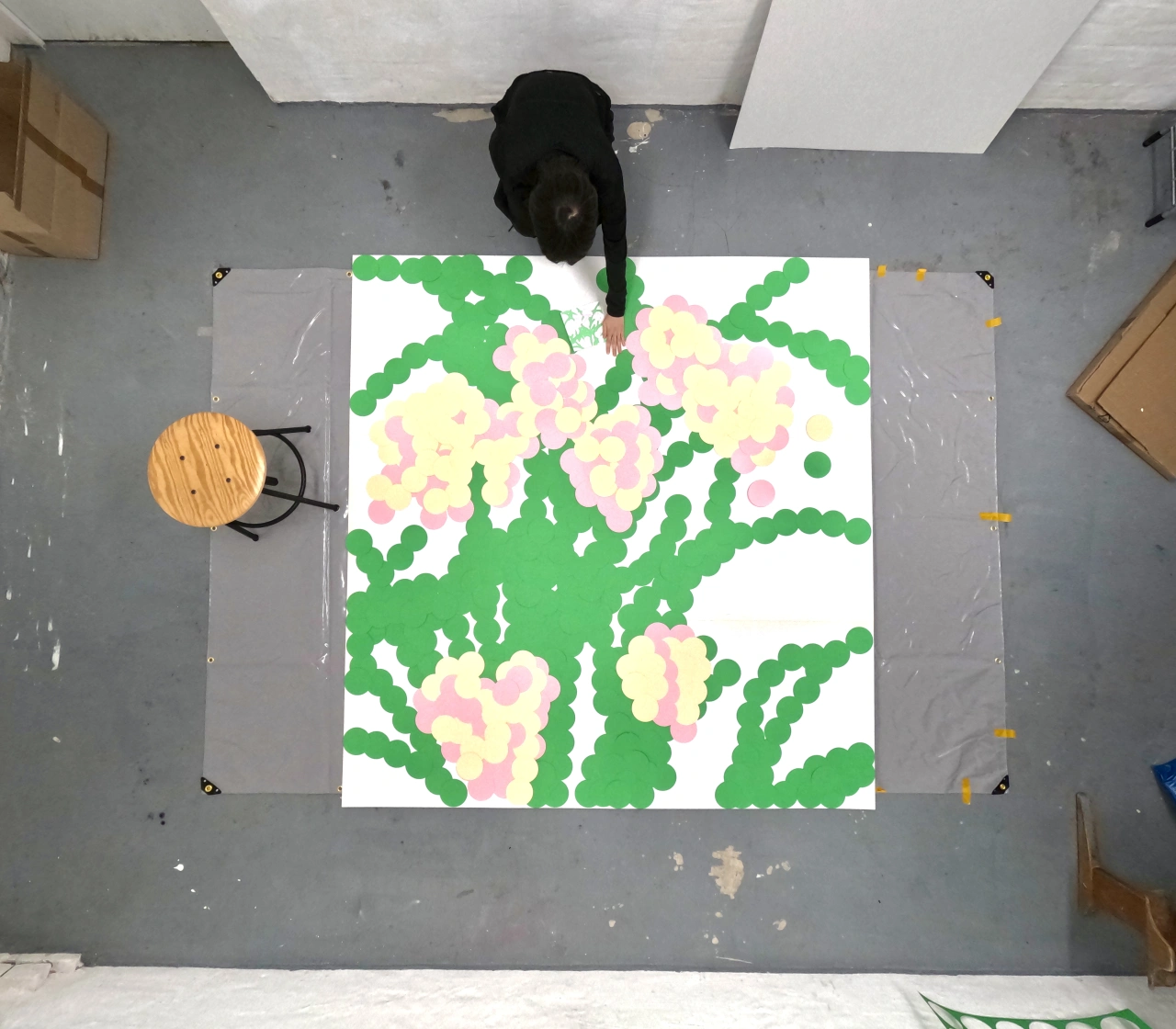Overhead view of artist Ayong Son standing beside a large hand-cut paper artwork featuring green cactus shapes with pink and cream flowers on a grey studio floor