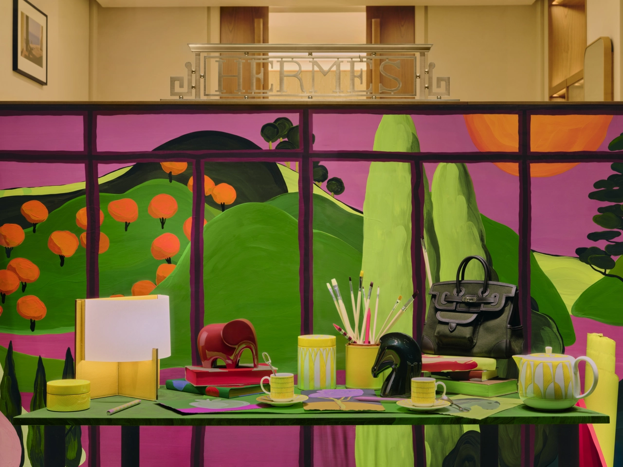 Colourful illustrated window display featuring a green shelf with yellow accessories, a black handbag, red bowl and potted plants against a vibrant pink and green background with orange flowers