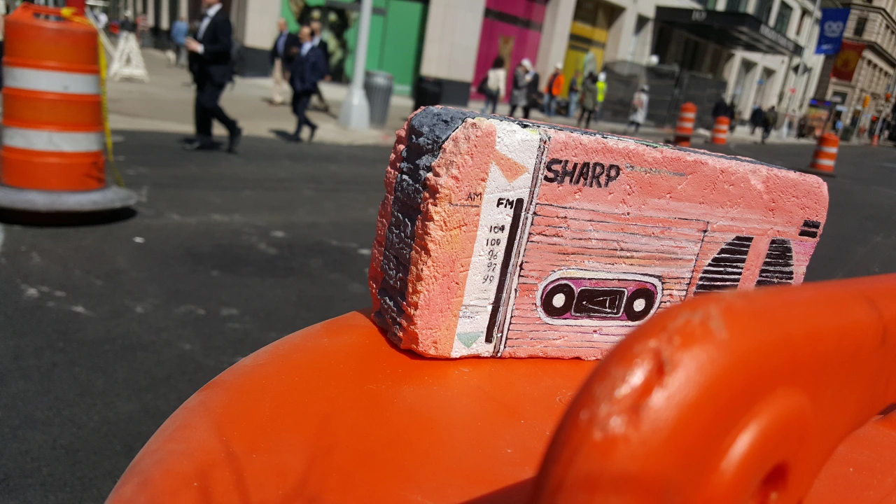 A painted rock depicting a coral-pink Sharp boombox cassette player, placed on an orange sculptural form on a city street