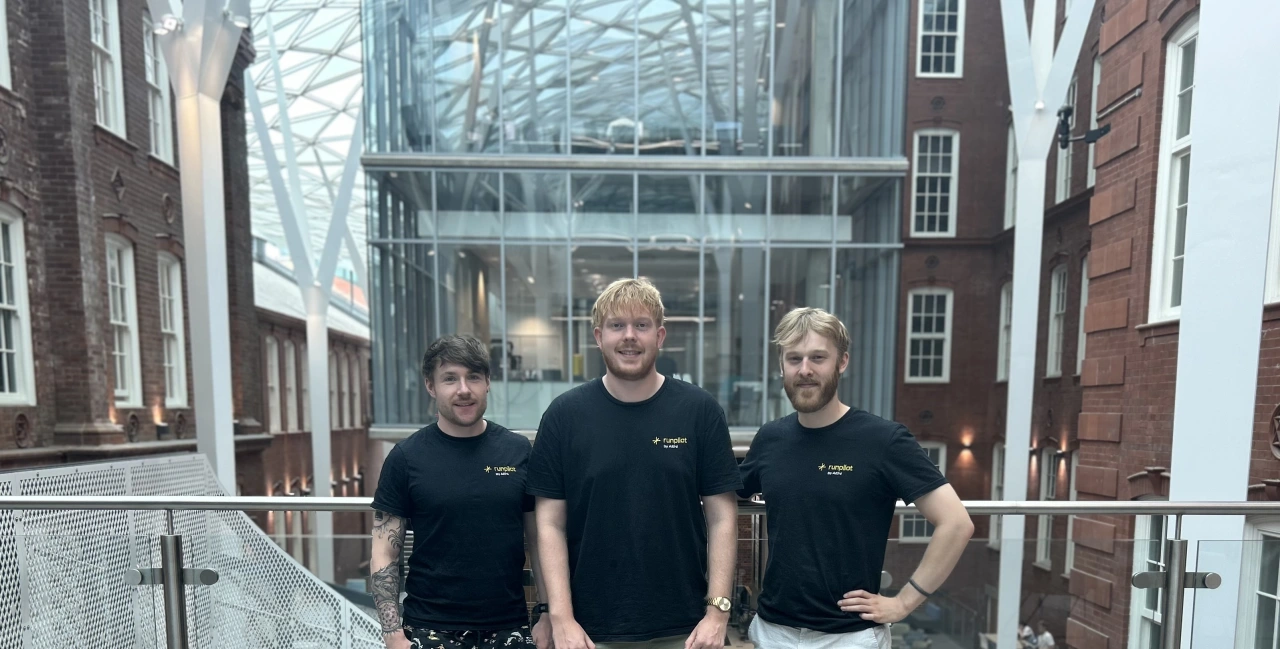Three men in black branded t-shirts stand on a modern glass bridge between brick buildings