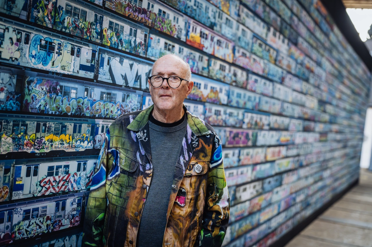 Man wearing glasses and a colourful jacket stands in front of a wall covered with colourful graffiti artworks and photographs
