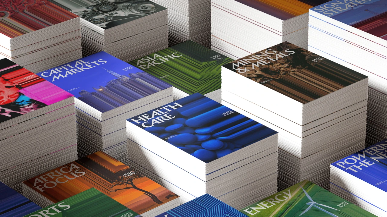 Stacks of printed materials with colorful covers displaying White & Case's new branding, featuring Catalyst Blue and kinetic typography against varied background colours