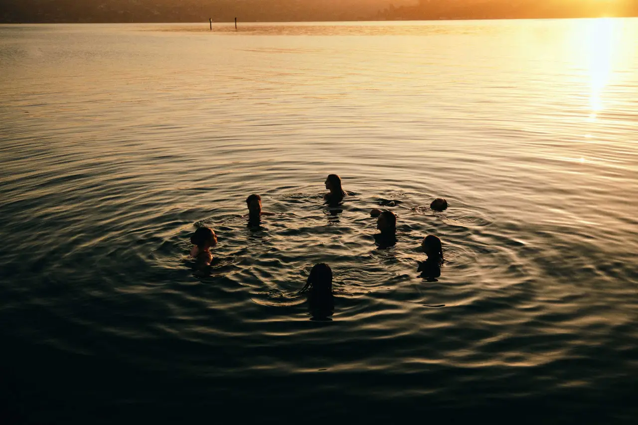Silhouetted figures in a circle in calm water at sunset, creating ripples around them