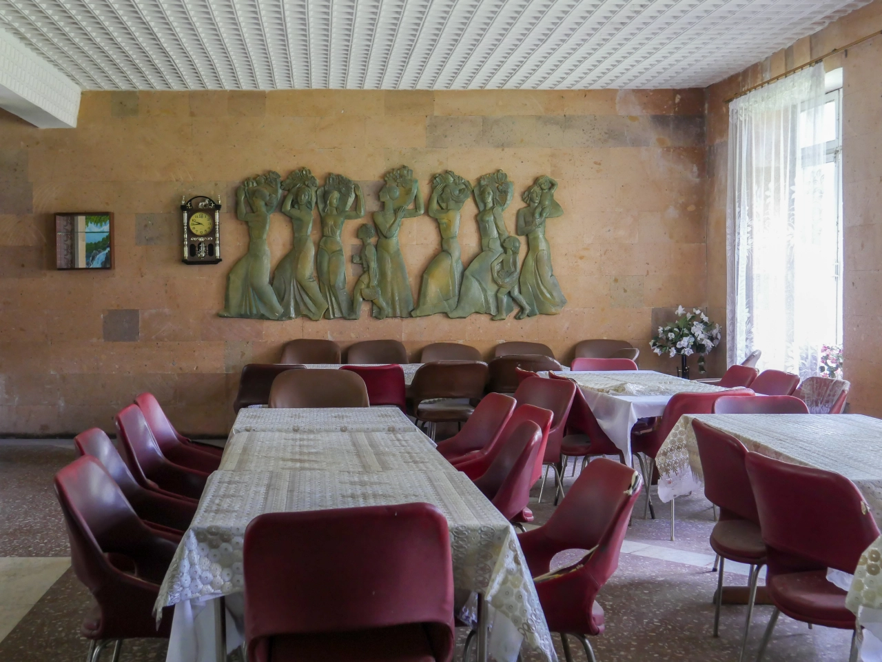 Dining room with terracotta walls, burgundy chairs, tables with white cloths, and a large green sculptural relief of figures above
