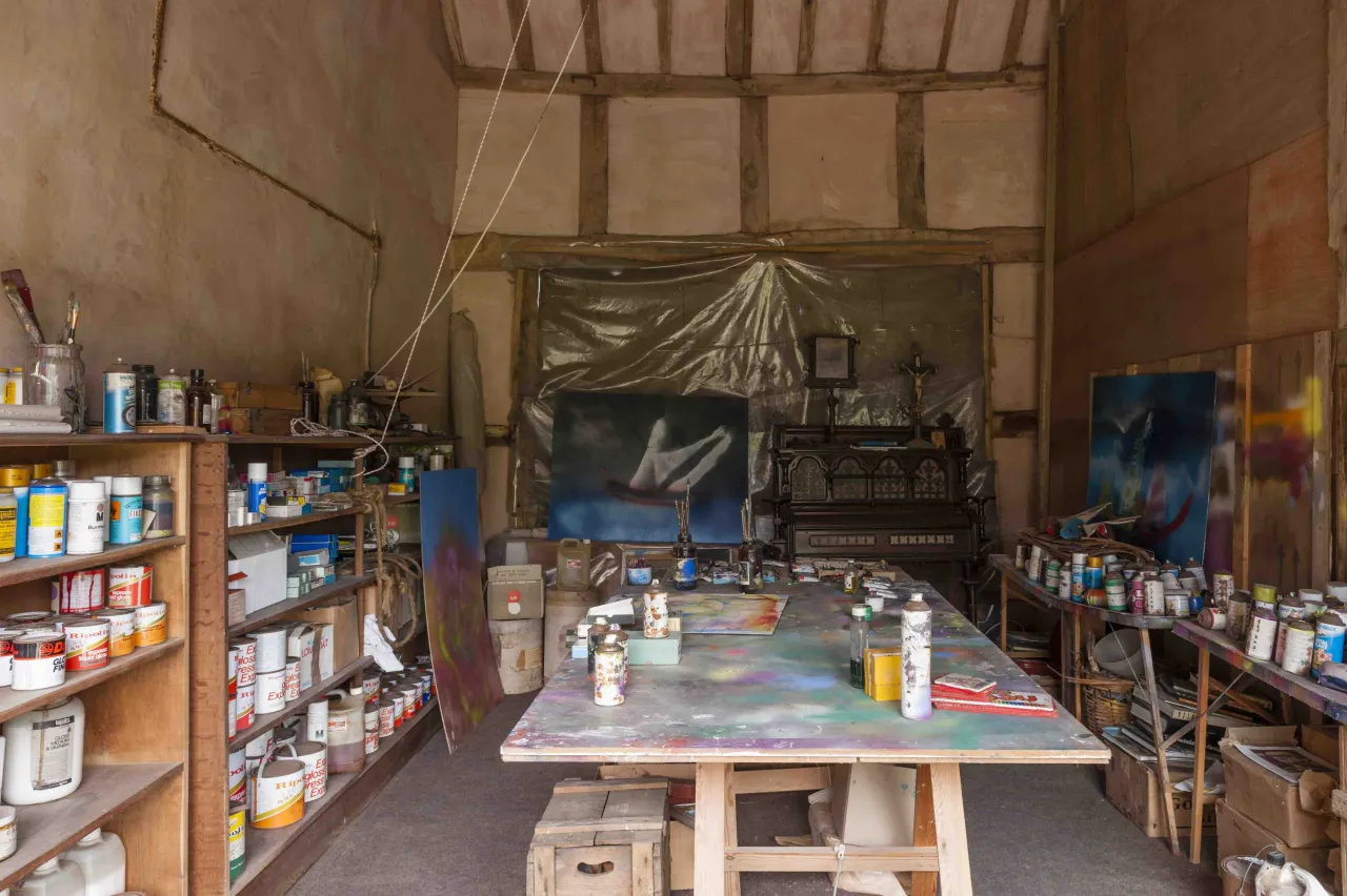 Sidney Nolan's studio at The Rodd, showing workbench with paint containers, shelves stacked with materials and cans, and covered furniture beneath plastic sheeting