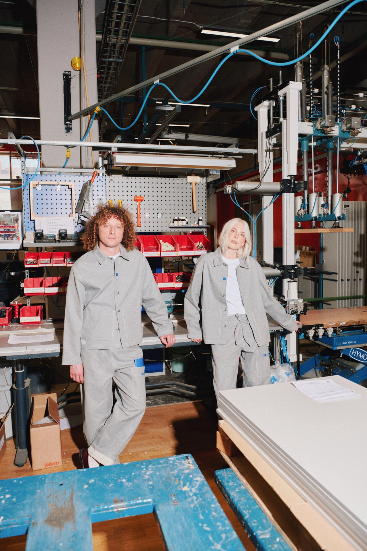 Two people wearing grey workwear uniforms stand in a bright workshop filled with tools, machinery and red storage bins