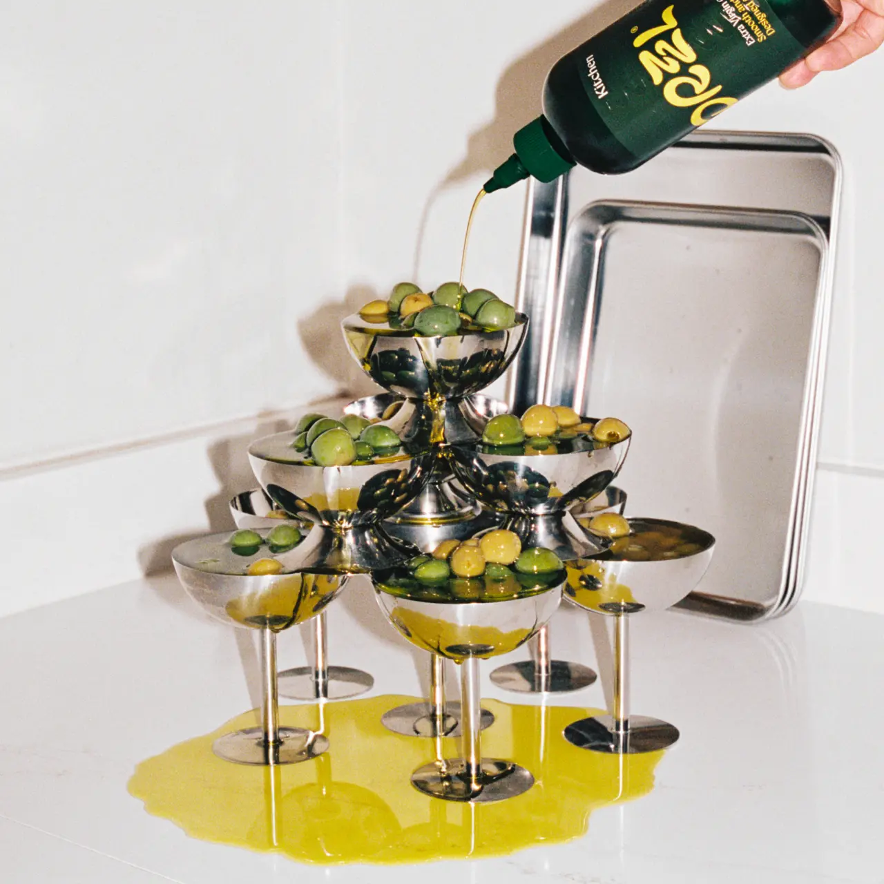 Drzzl olive oil pouring from a bottle onto a tiered gold stand filled with olives and martini glasses, with a napkin dispenser beside it