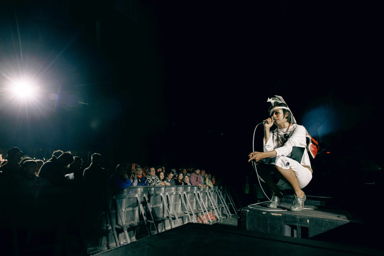A performer in a unique white costume and headpiece crouches near the edge of a stage with a microphone, singing to a crowd under spotlight illumination.