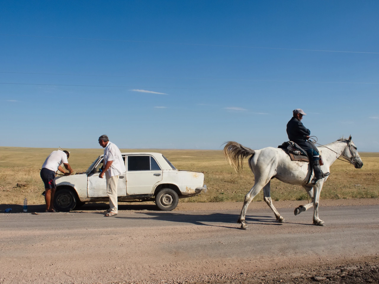 A man on horseback passes a white car broken down on a steppe road, with two people attending to the vehicle under a clear blue sky