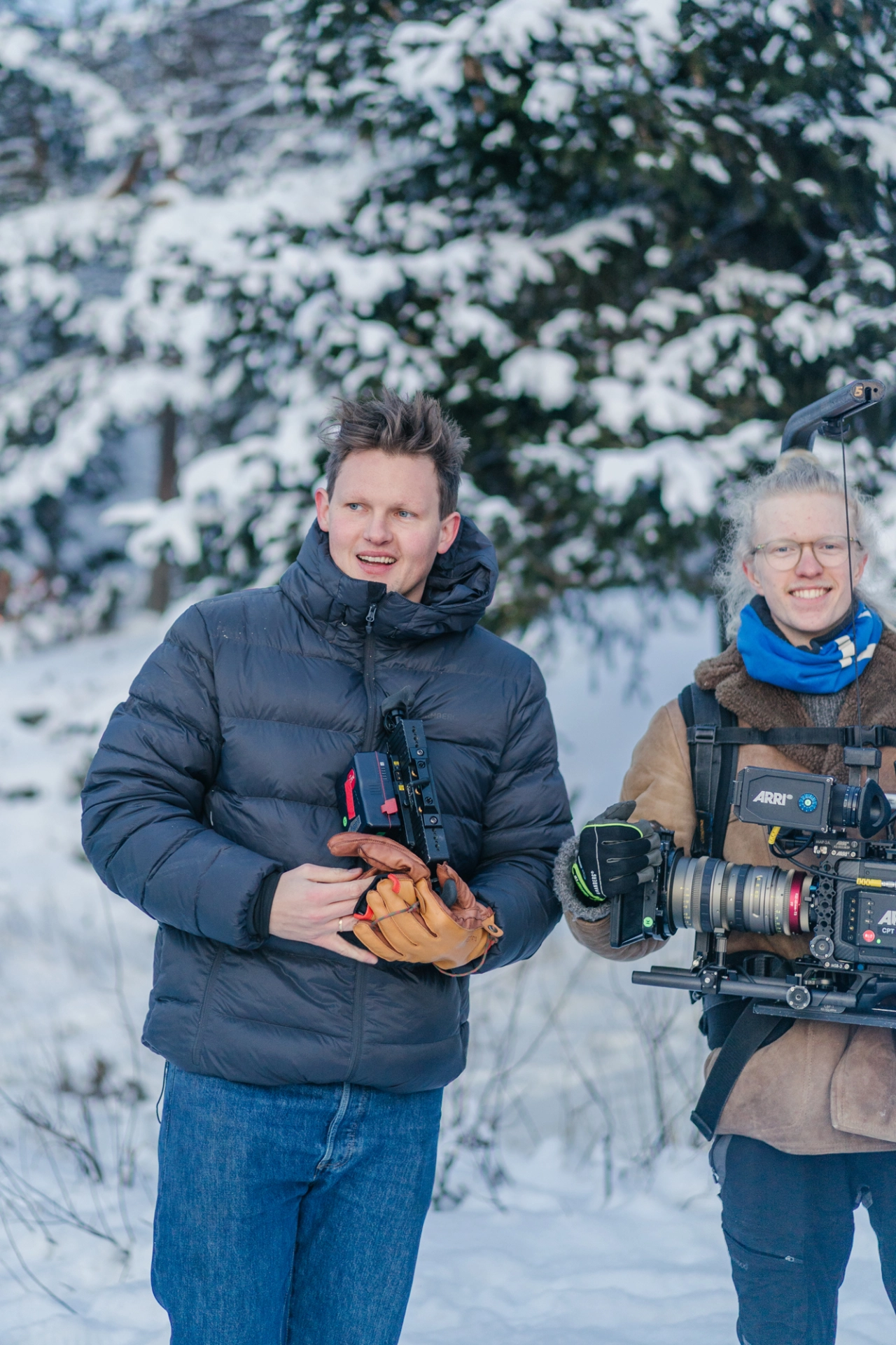 Behind-the-scenes crew member operating film camera alongside an actor in winter clothing against snowy forest backdrop