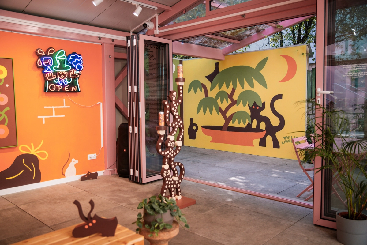 Interior of Philip Lindeman's exhibition showing an orange wall with neon sign and illustration, yellow wall with cat and tree mural, wooden sculptures and potted plant on the floor, and pink-framed windows and doors.