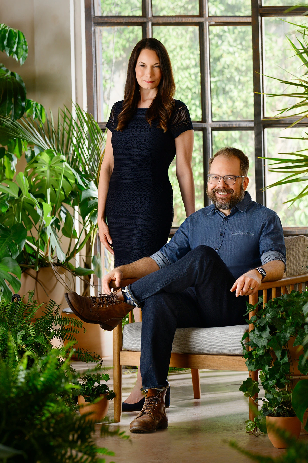 Jennifer Spire and Dustin Black stand among potted plants in front of a large gridded window in an interior space