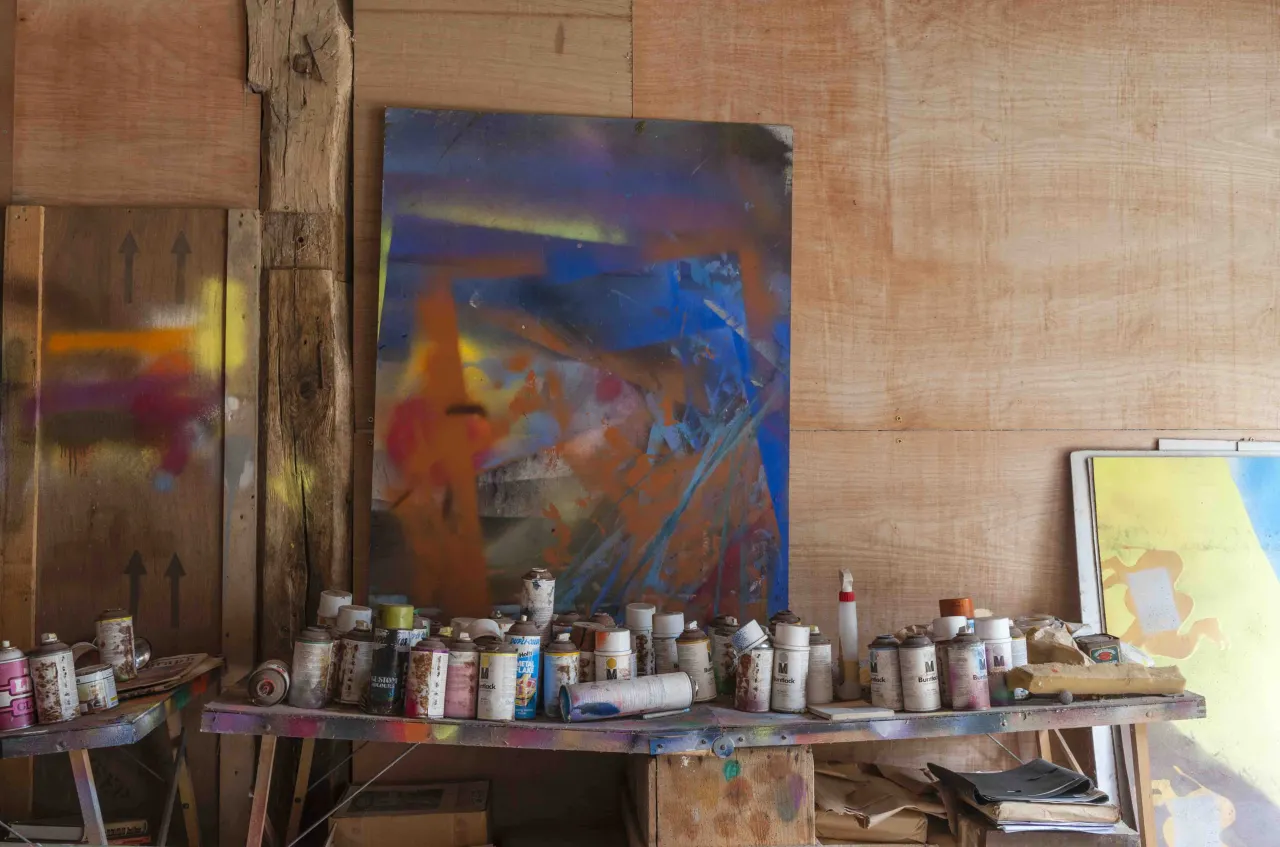 Rows of paint cans and spray paint bottles arranged on a wooden shelf beneath a colourful abstract painting in Sidney Nolan's studio at The Rodd