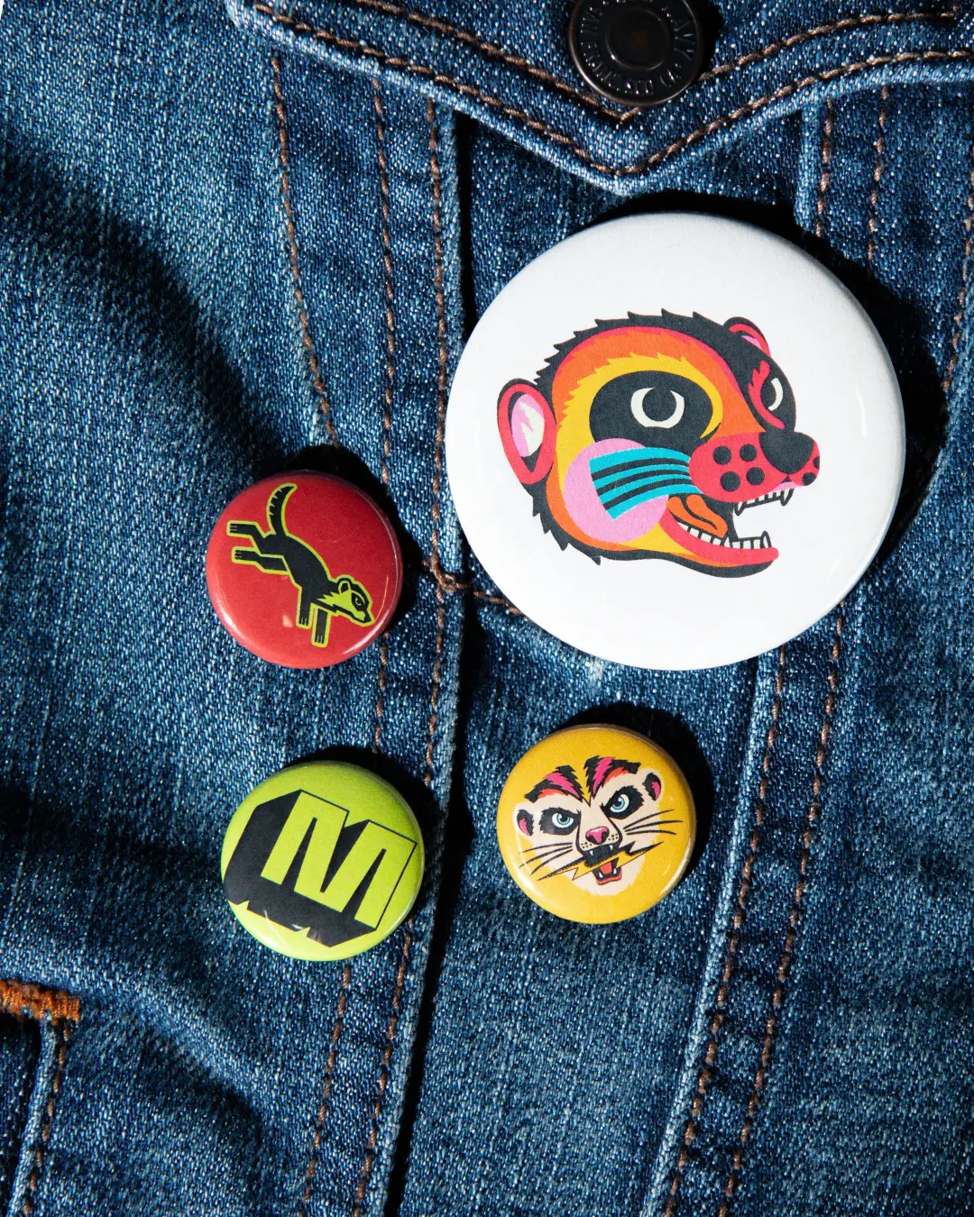 Four colourful enamel pin badges featuring a meerkat character design stuck to denim fabric
