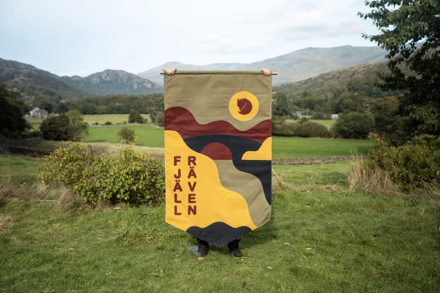 Fjällräven banner hand-stitched by Miscellaneous Adventures hanging in a Lake District valley, featuring geometric shapes and the text 'Fjällräven Classic' in yellow and burgundy tones against mountains and green fields