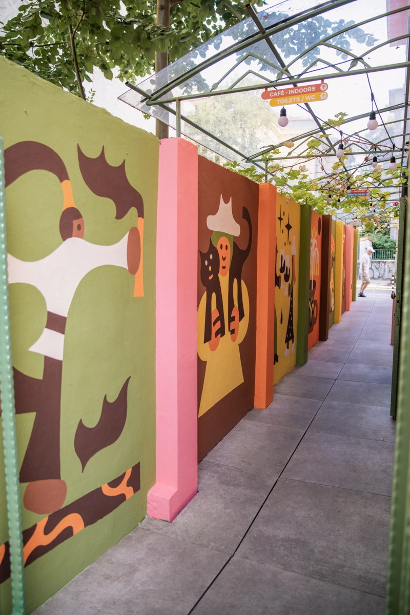 A covered corridor displays colourful illustrated panels featuring stylised cats and figures in warm tones of yellow, orange and brown against green and pink backgrounds