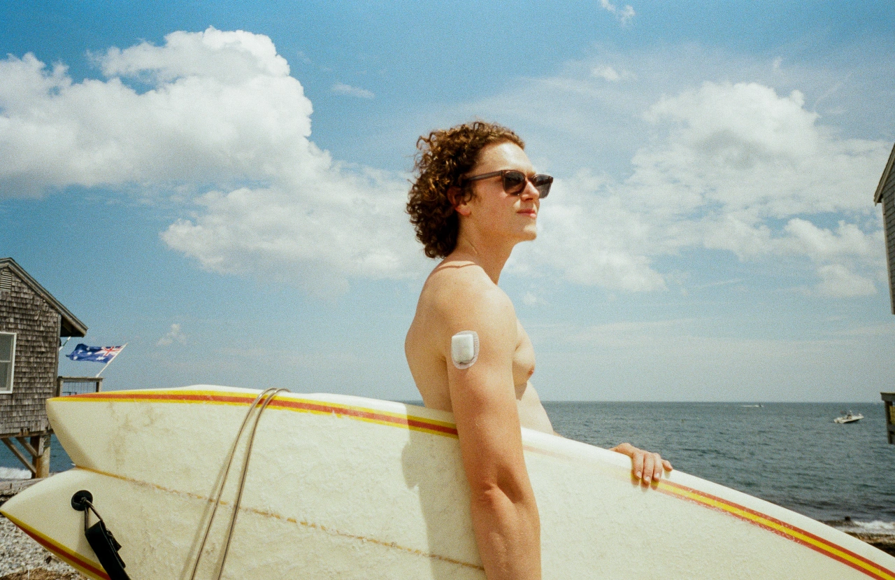Shirtless man wearing sunglasses holding a surfboard on a beach, with a sun protection sticker visible on his arm and the sea behind him