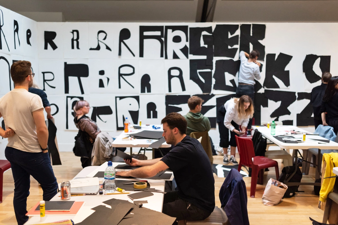 Participants engage in collaborative workshop activities around tables with a large black and white typographic design installation on the wall behind them at International Assembly