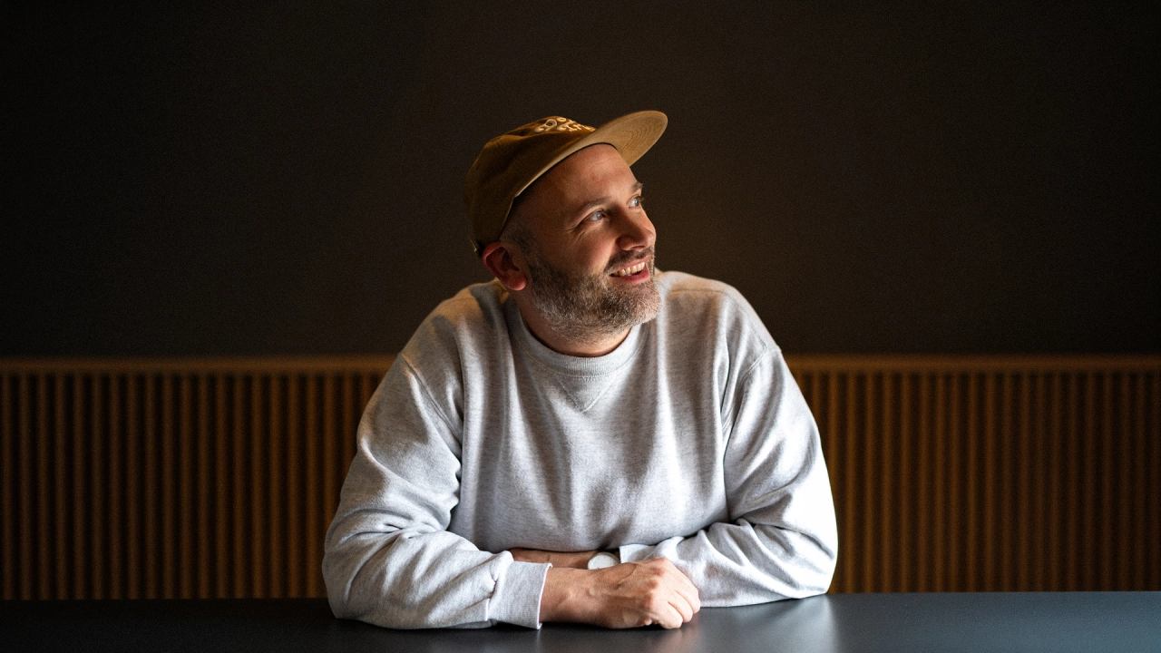 Jeff Bowerman, smiling man wearing a light grey sweatshirt and tan cap, seated at a table against a dark wall with wooden panelling