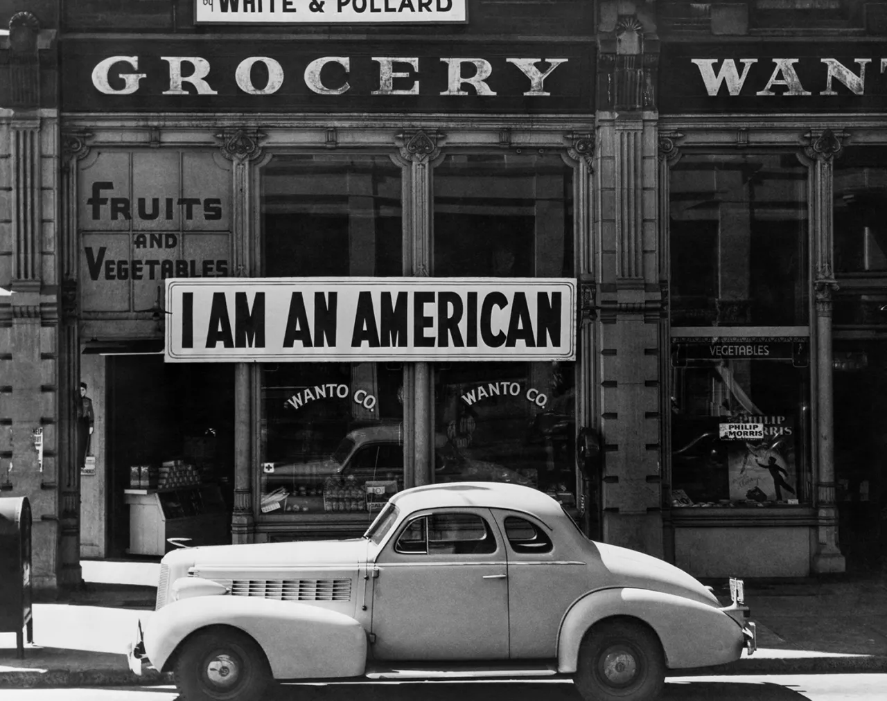 Black and white photograph of a 1940s car parked outside a grocery store storefront bearing a large sign reading "I AM AN AMERICAN