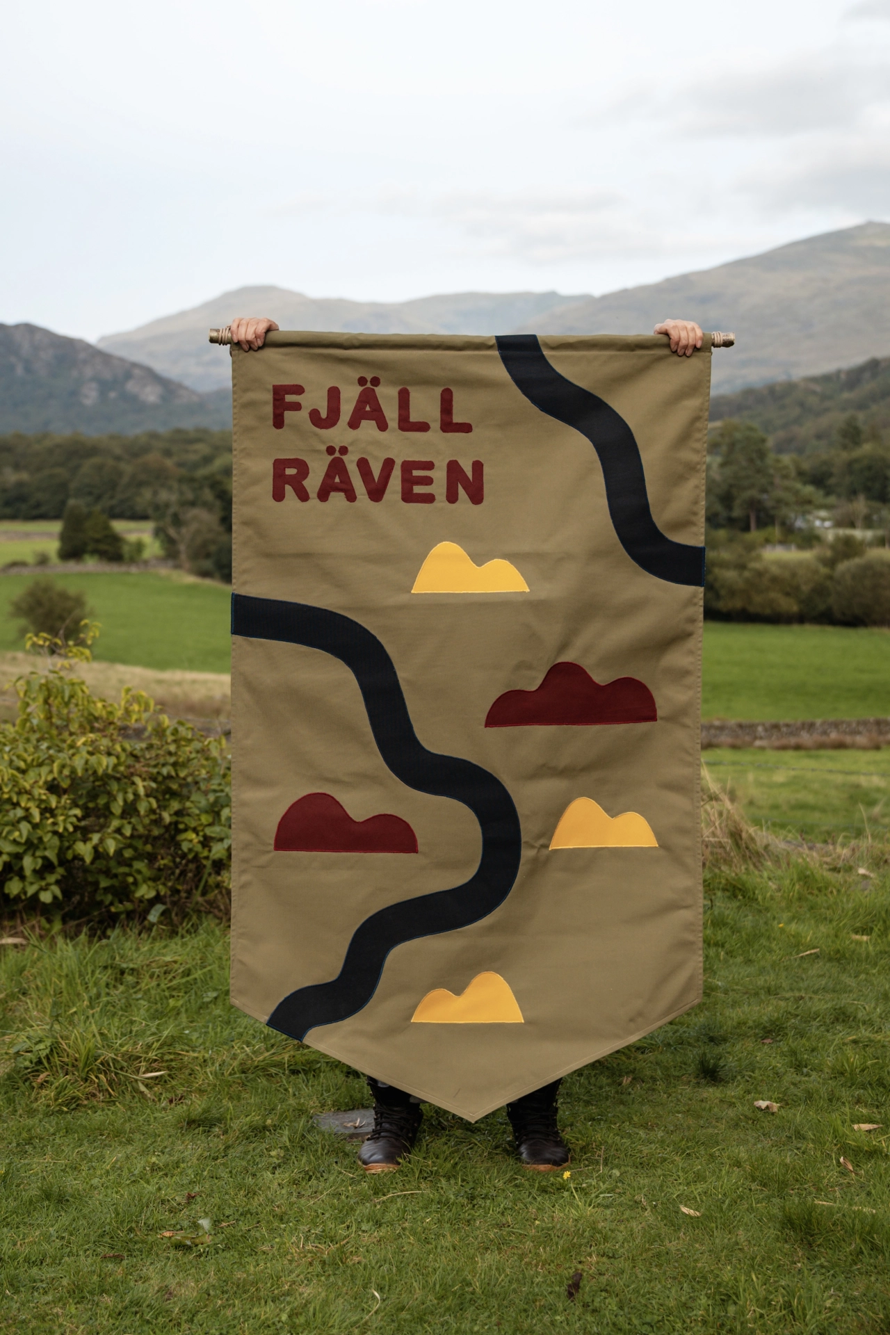 Tan banner with "FJÄLL RÄVEN" text and geometric mountain shapes in yellow, burgundy and black, held up outdoors with mountains visible in the background