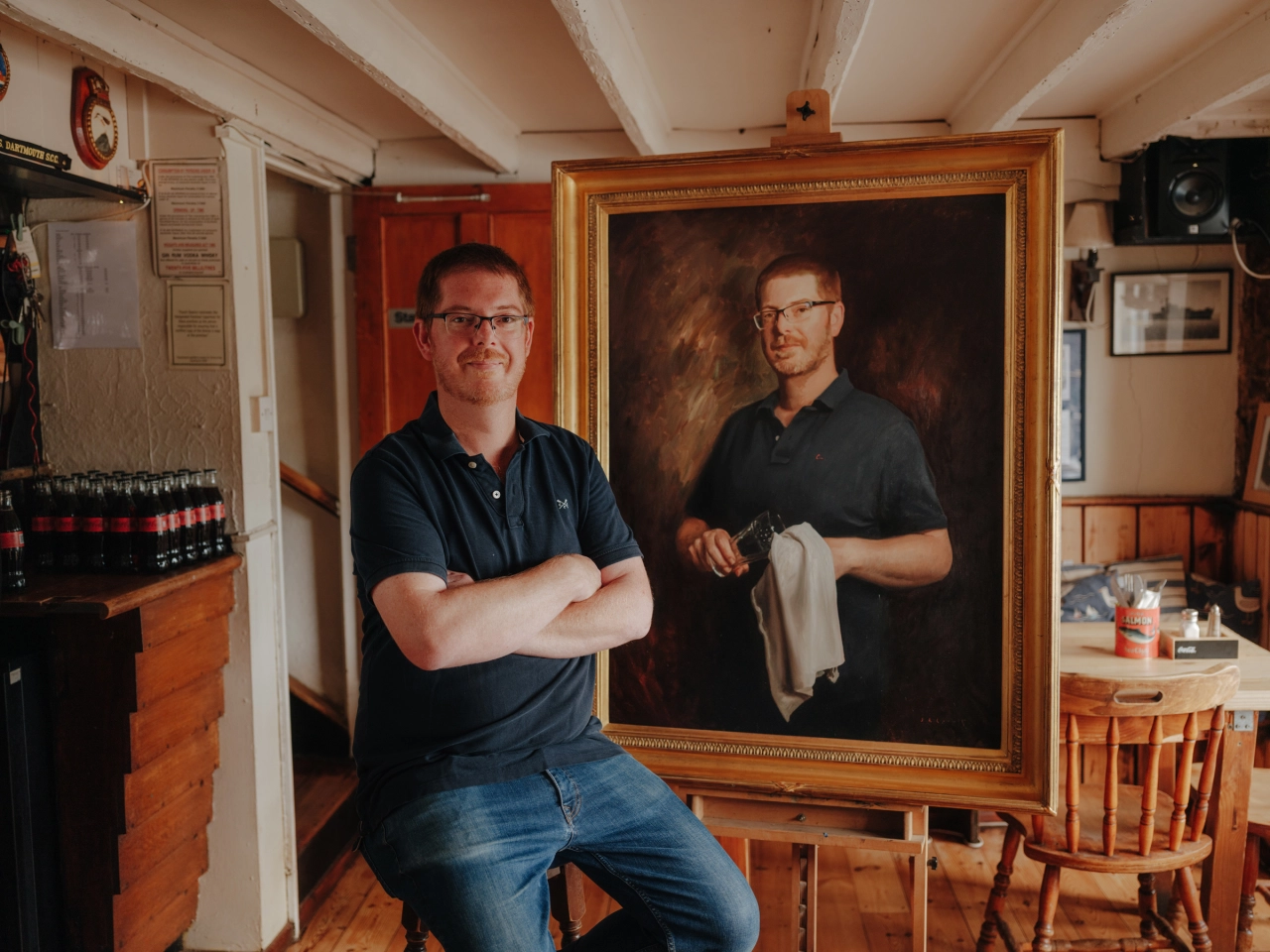 Man in dark polo shirt and glasses standing with arms crossed beside a large gilt-framed oil portrait of himself in a studio setting.