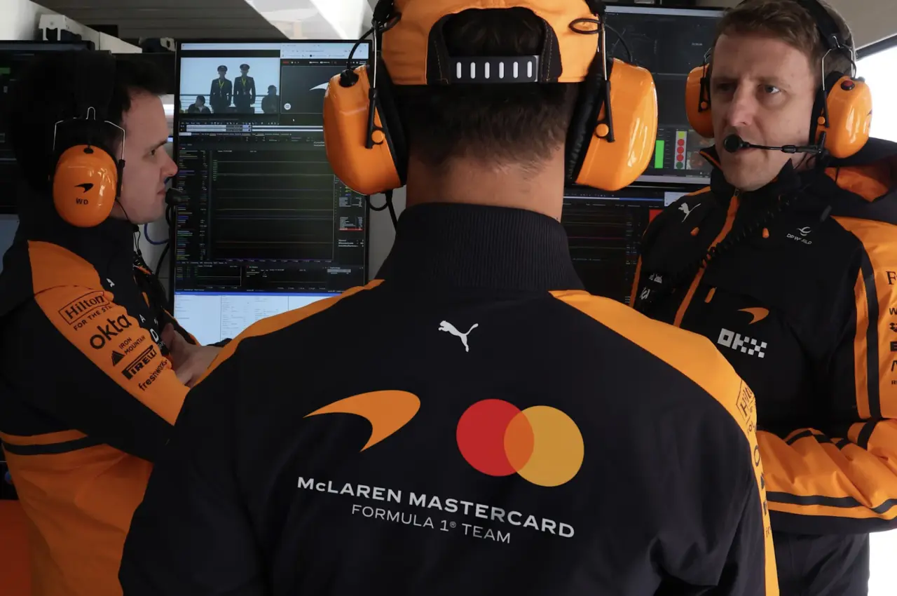 Three McLaren Formula 1 team members wearing orange and black team wear and headphones work at a monitor displaying video editing software in a garage setting.