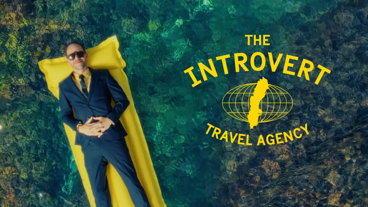 Man in business suit and sunglasses lying on yellow raft with "The Introvert Travel Agency" logo and globe graphic on teal water background