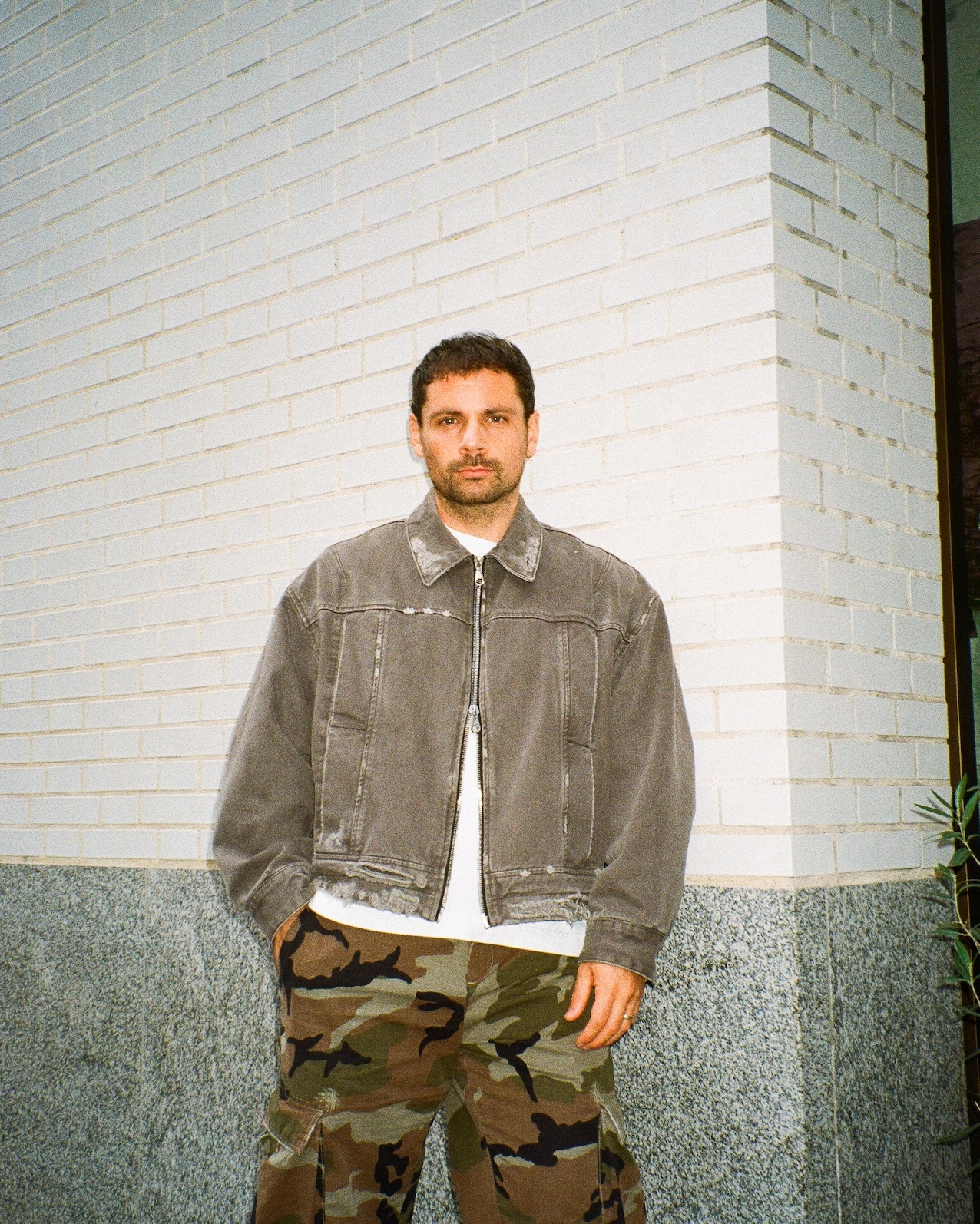 Conall McAteer stands against a white brick wall wearing a grey denim jacket over camouflage trousers