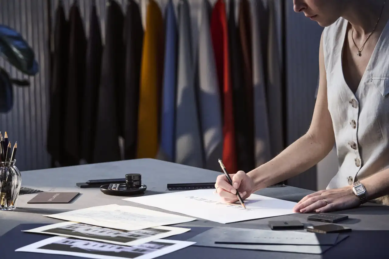 Designer sketching on white paper at a desk with fabric swatches hanging on a rail behind her
