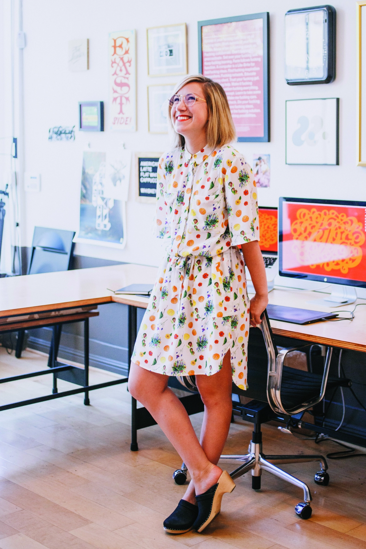 Jessica Hische wearing a colourful fruit and vegetable patterned dress standing in a creative studio with artwork and a computer monitor visible on the wall behind her