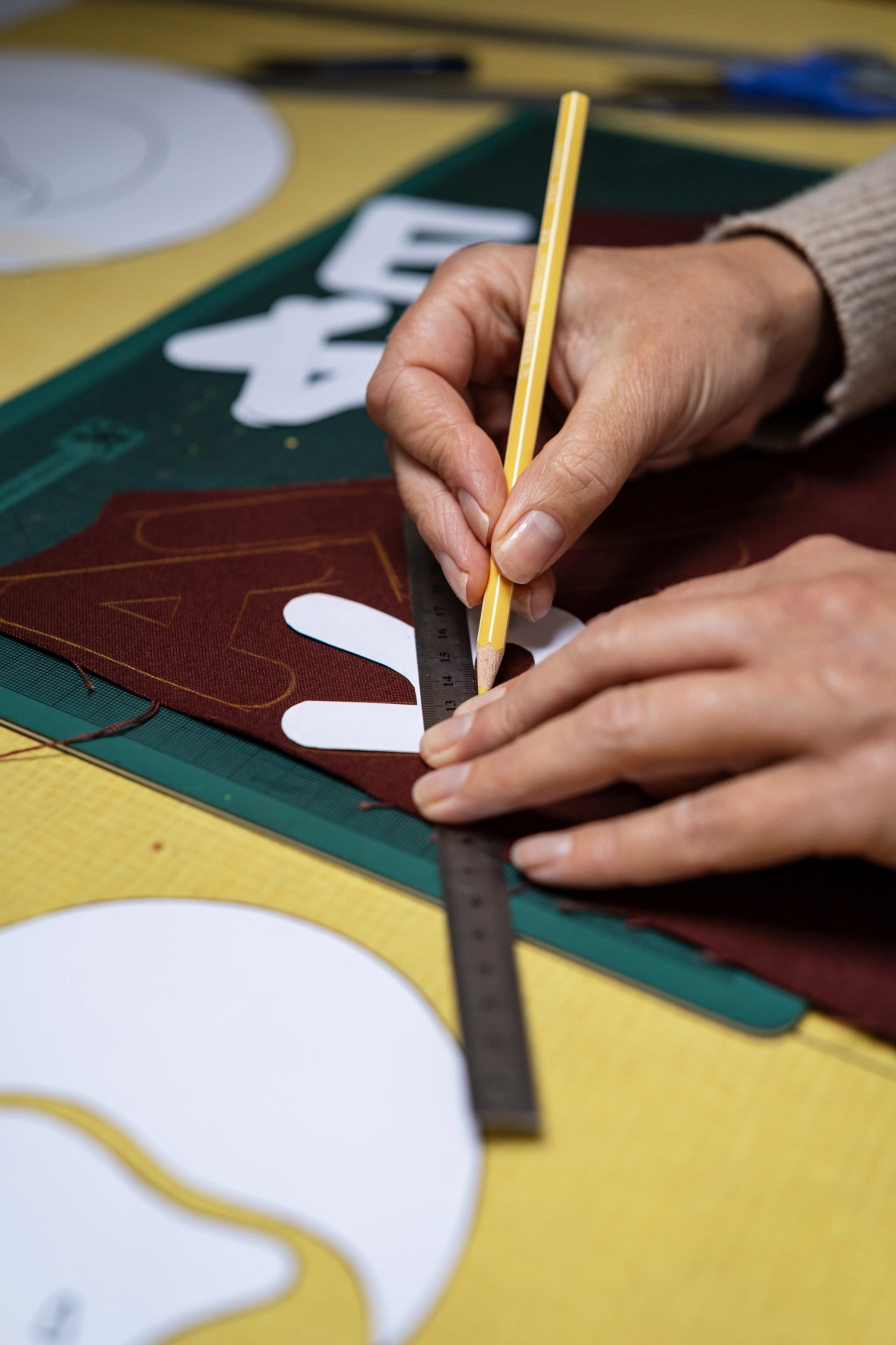 Hands holding a yellow pencil and ruler while sketching white curved shapes onto burgundy and green fabric sections of a handmade banner