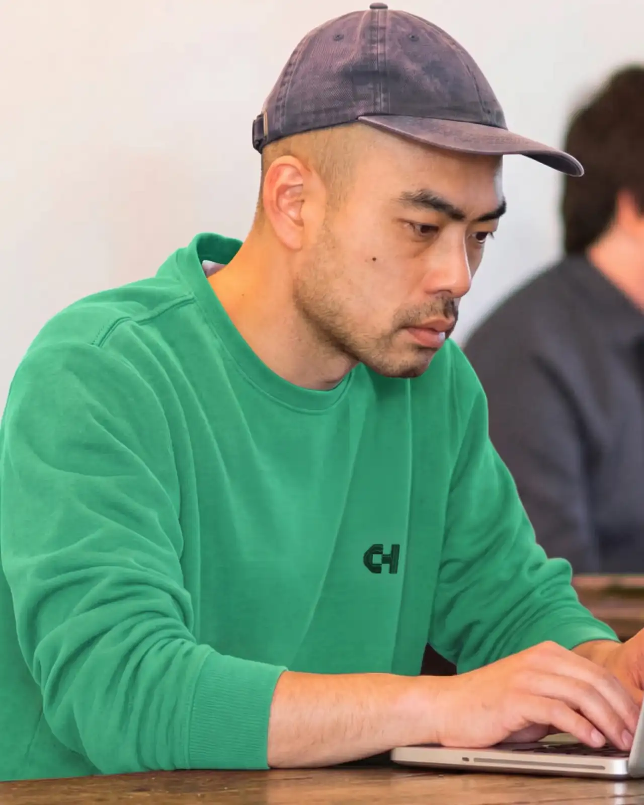 Man in green hoodie with CH logo wearing dark cap, working at laptop