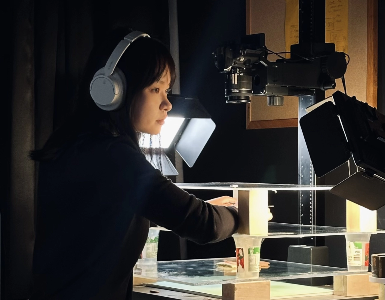Animator Qiaoyu Liao wearing headphones sits at a multiplane camera rig with illuminated desk, recording voice or sound for the animation project