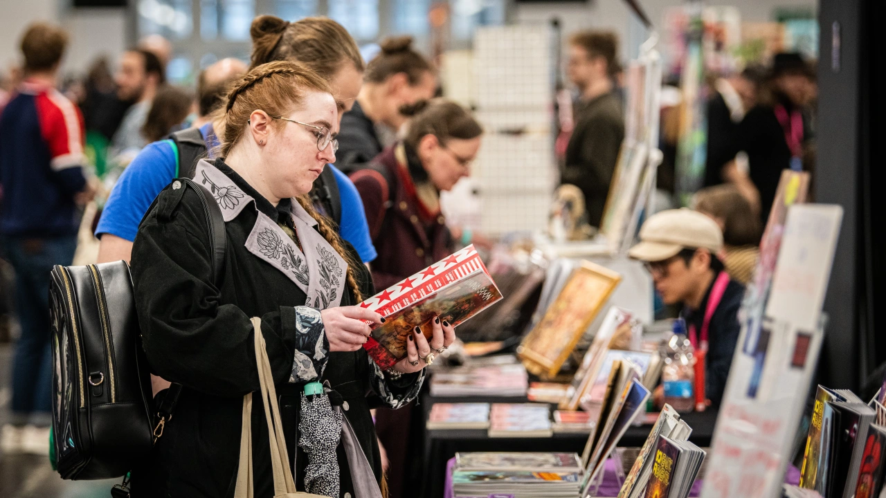 A visitor with glasses and a backpack browses comic books and graphic novels at a crowded convention stall during Thought Bubble festival