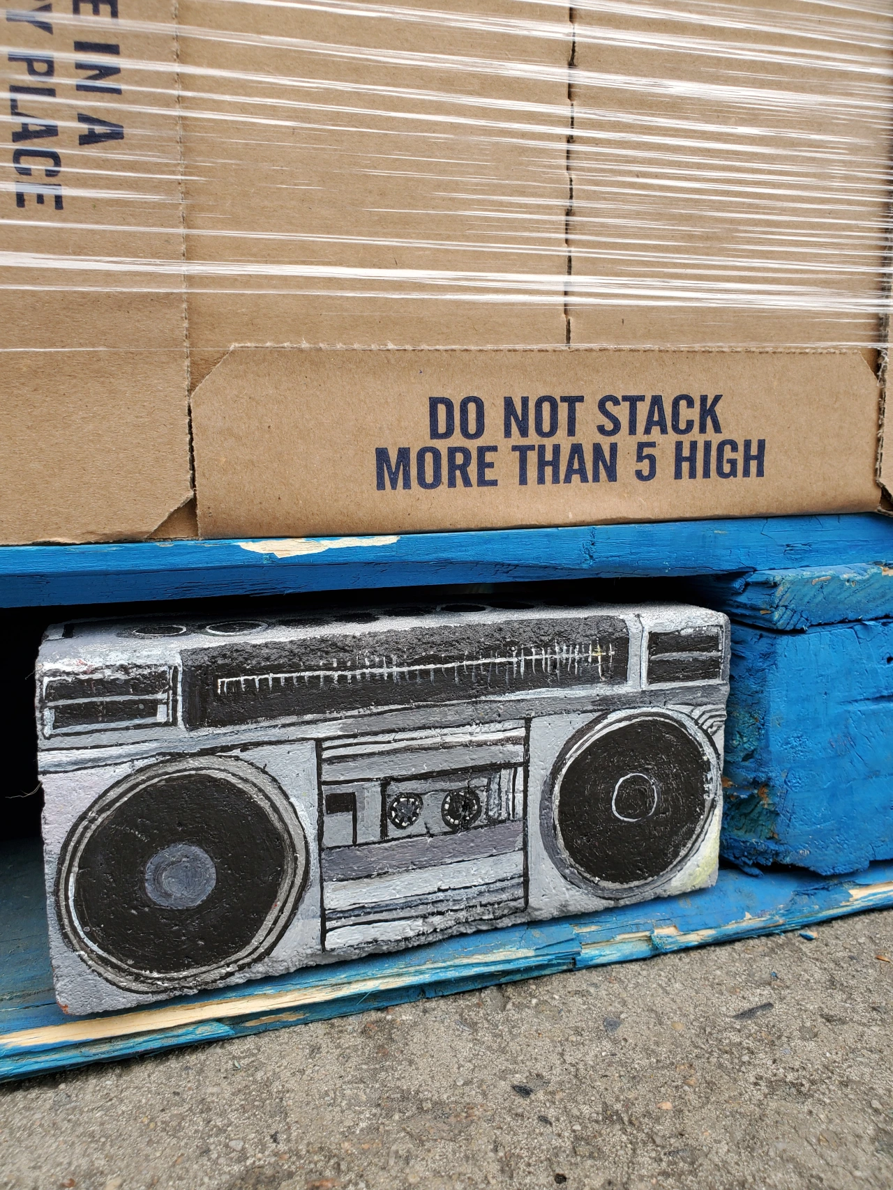 Stacked cardboard boxes with a printed boombox artwork on a blue wooden pallet, with shipping label reading "DO NOT STACK MORE THAN 5 HIGH